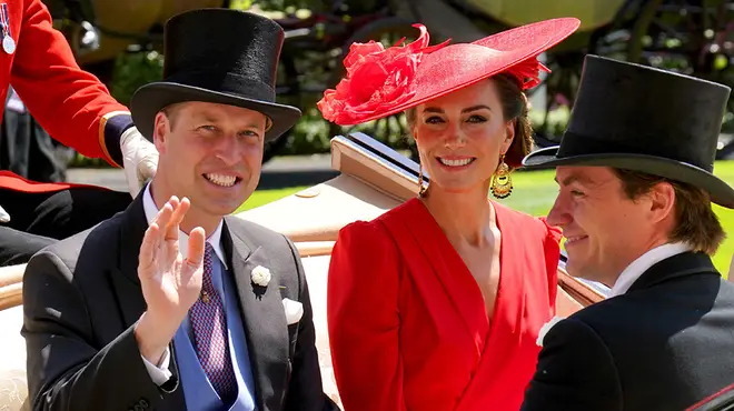 Prince William and Kate Middleton at the races in a carriage. Kate is wearing a red dress and hat and William is smiling and waving while wearing a top hat