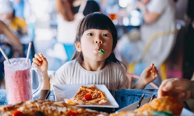 Young child with food in her mouth and a pizza in front of her in a restaurant