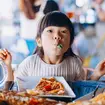 Young child with food in her mouth and a pizza in front of her in a restaurant